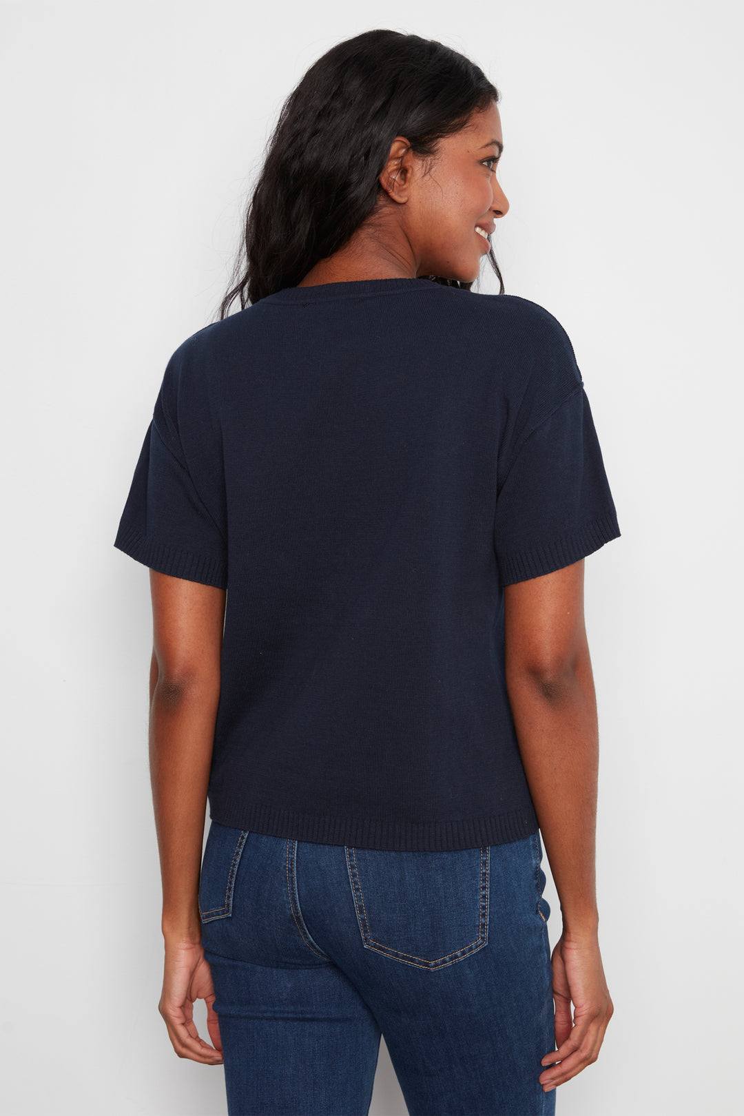 A woman shown from the back wearing a dark navy blue short-sleeve knit top and blue jeans against a plain light gray background.