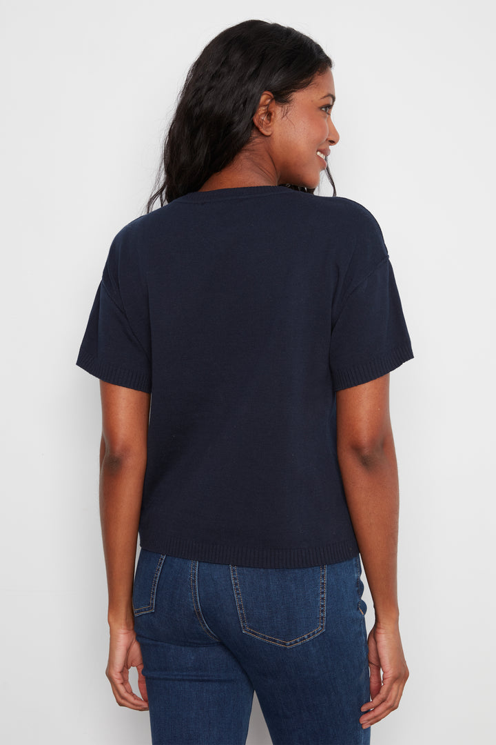A woman shown from the back wearing a dark navy blue short-sleeve knit top and blue jeans against a plain light gray background.