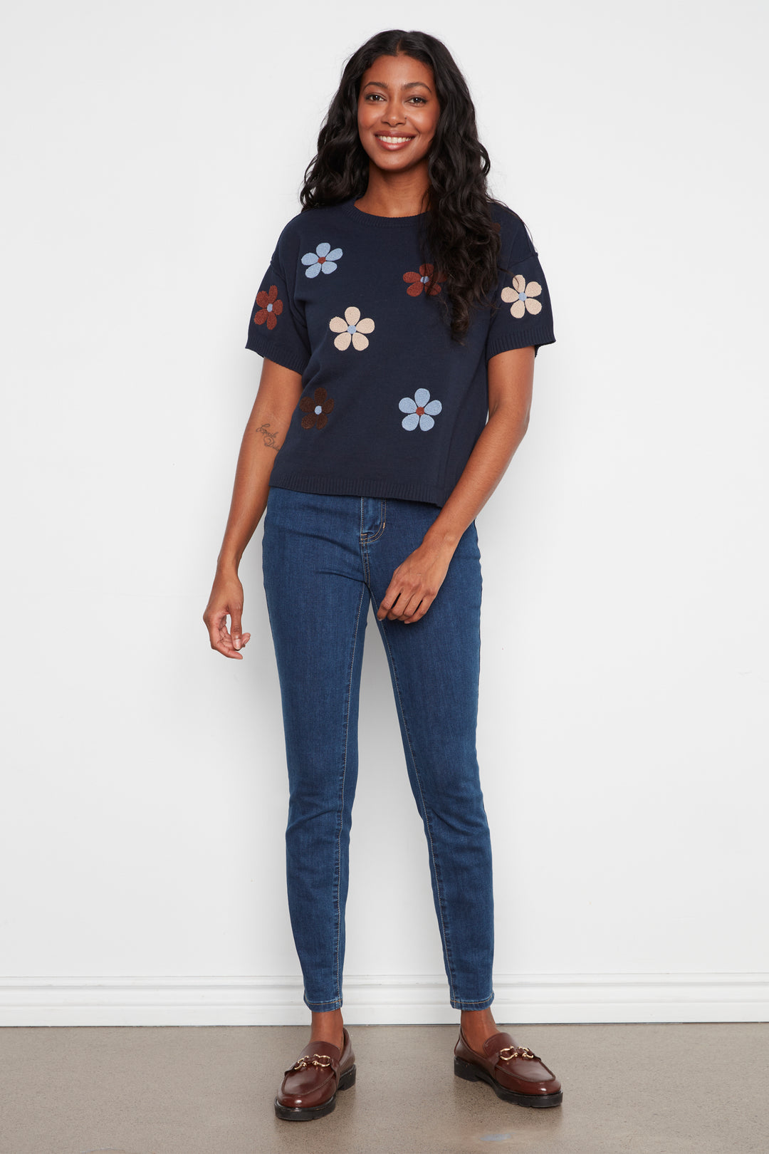 A woman modeling a navy blue short-sleeve t-shirt with colorful embroidered flowers, paired with blue jeans and brown loafers, standing against a plain white wall.