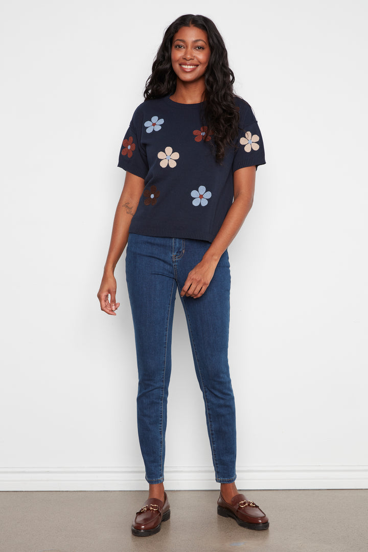 A woman modeling a navy blue short-sleeve t-shirt with colorful embroidered flowers, paired with blue jeans and brown loafers, standing against a plain white wall.