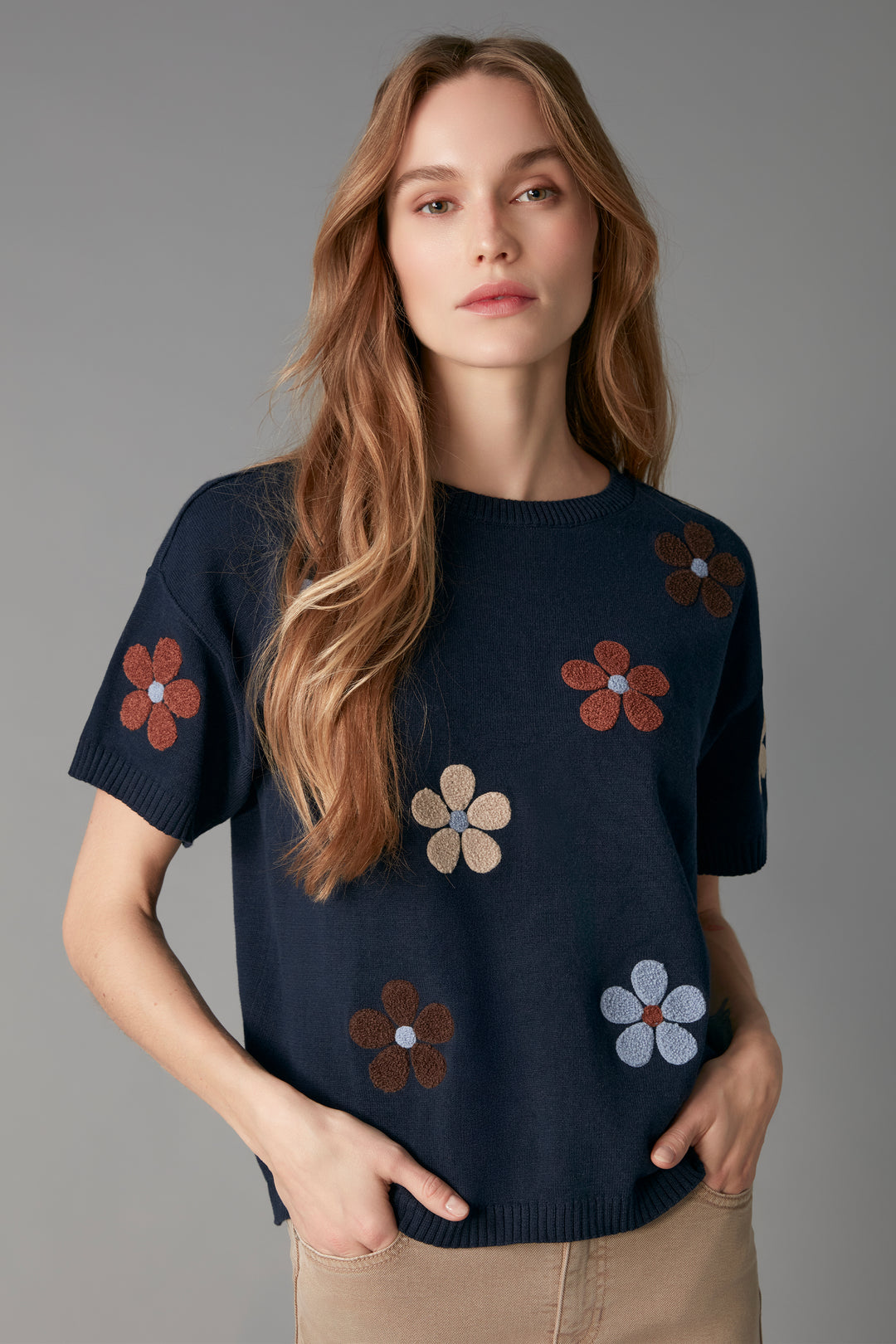 A woman modeling a navy blue short-sleeve knit sweater with embroidered flower motifs in brown, beige, and light blue, paired with beige pants against a solid gray studio background.