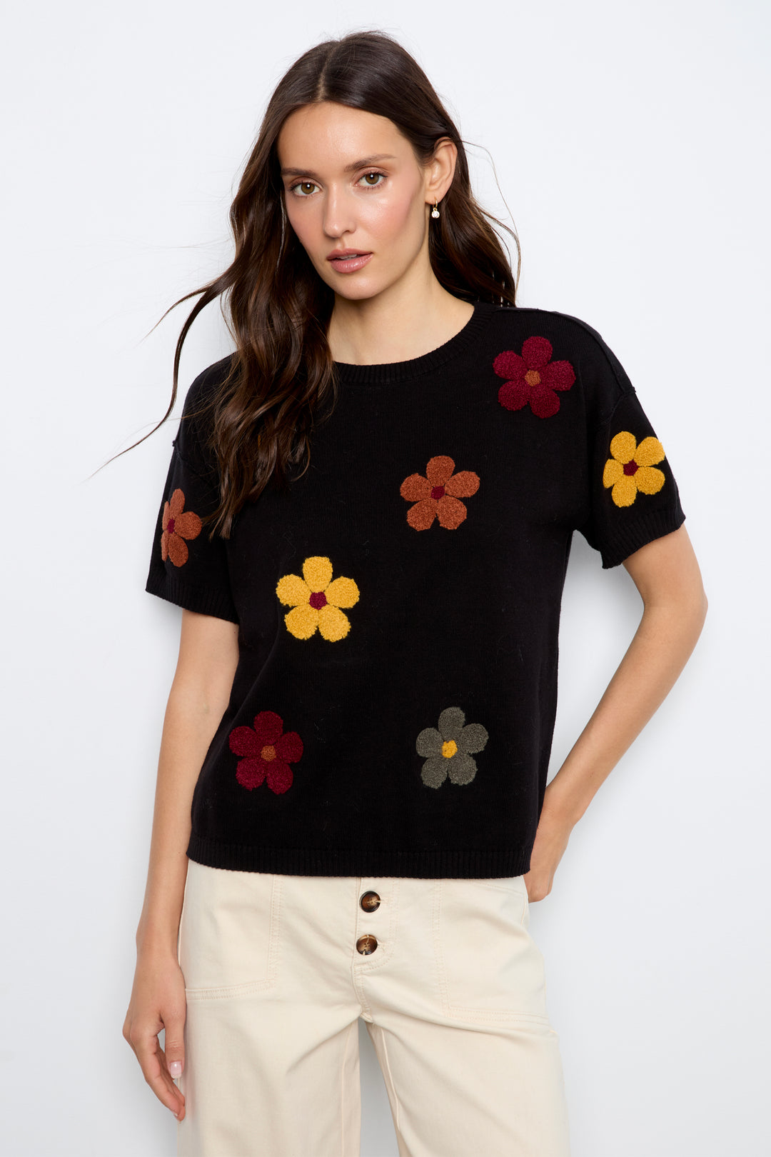 A woman modeling a black short-sleeve knit top with colorful embroidered flowers, paired with cream jeans, against a plain light gray background.