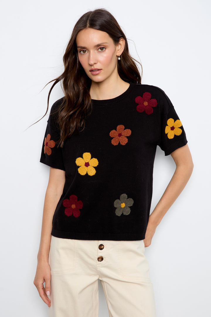 A woman modeling a black short-sleeve knit top with colorful embroidered flowers, paired with cream jeans, against a plain light gray background.