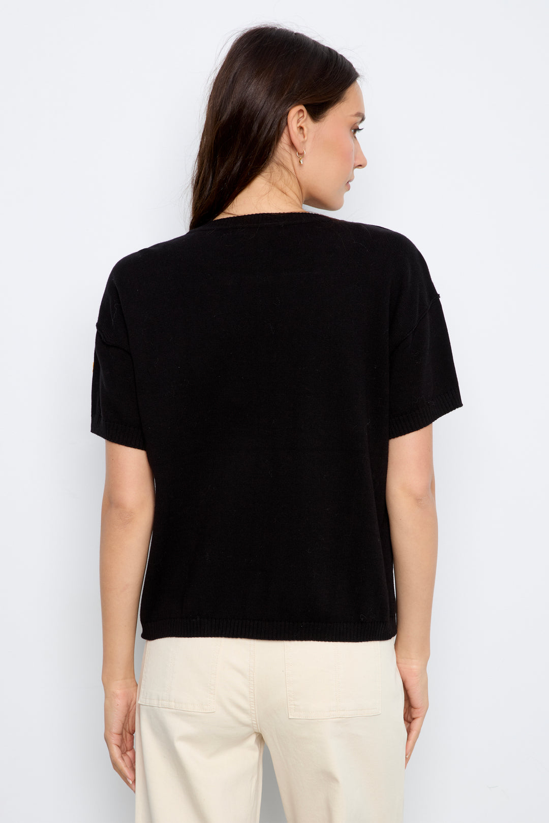 A woman shown from the back modeling a black short-sleeve knit top with colorful embroidered flowers, paired with cream jeans, against a plain light gray background.