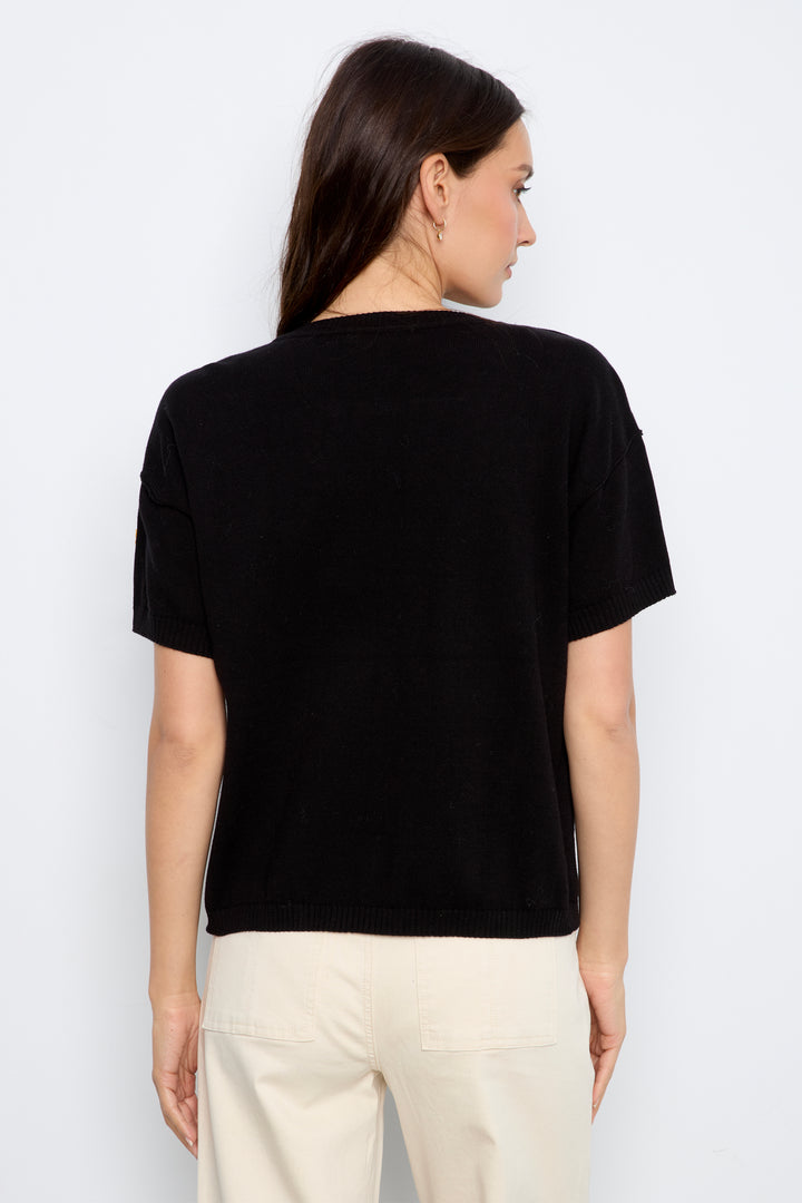 A woman shown from the back modeling a black short-sleeve knit top with colorful embroidered flowers, paired with cream jeans, against a plain light gray background.