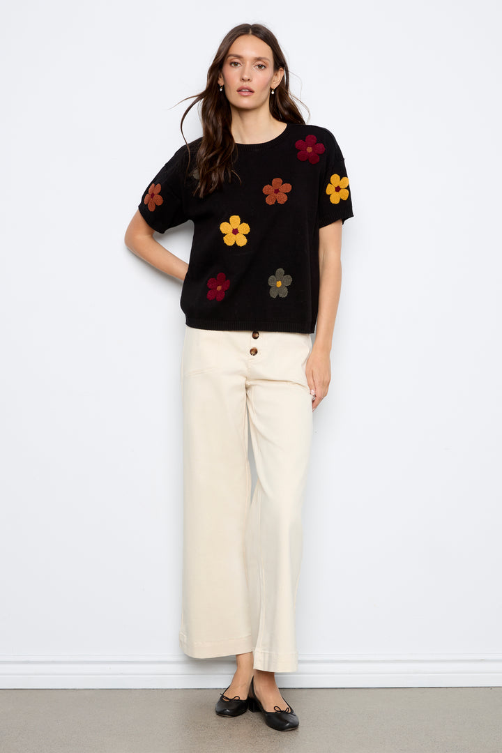 A woman modeling a black short-sleeve knit top with colorful embroidered flowers, paired with cream jeans, against a plain light gray background.