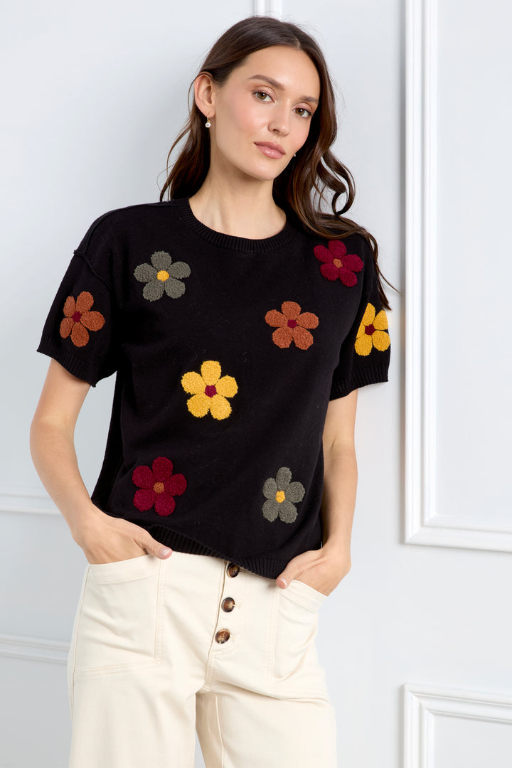 A woman modeling a black short-sleeve knit top with colorful embroidered flowers, paired with cream jeans, against a light gray background.