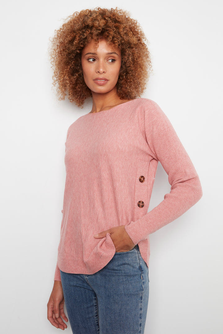 A woman with curly brown hair wearing a dusty pink long-sleeve knit sweater with brown buttons on the side, paired with blue jeans, against a plain white background.