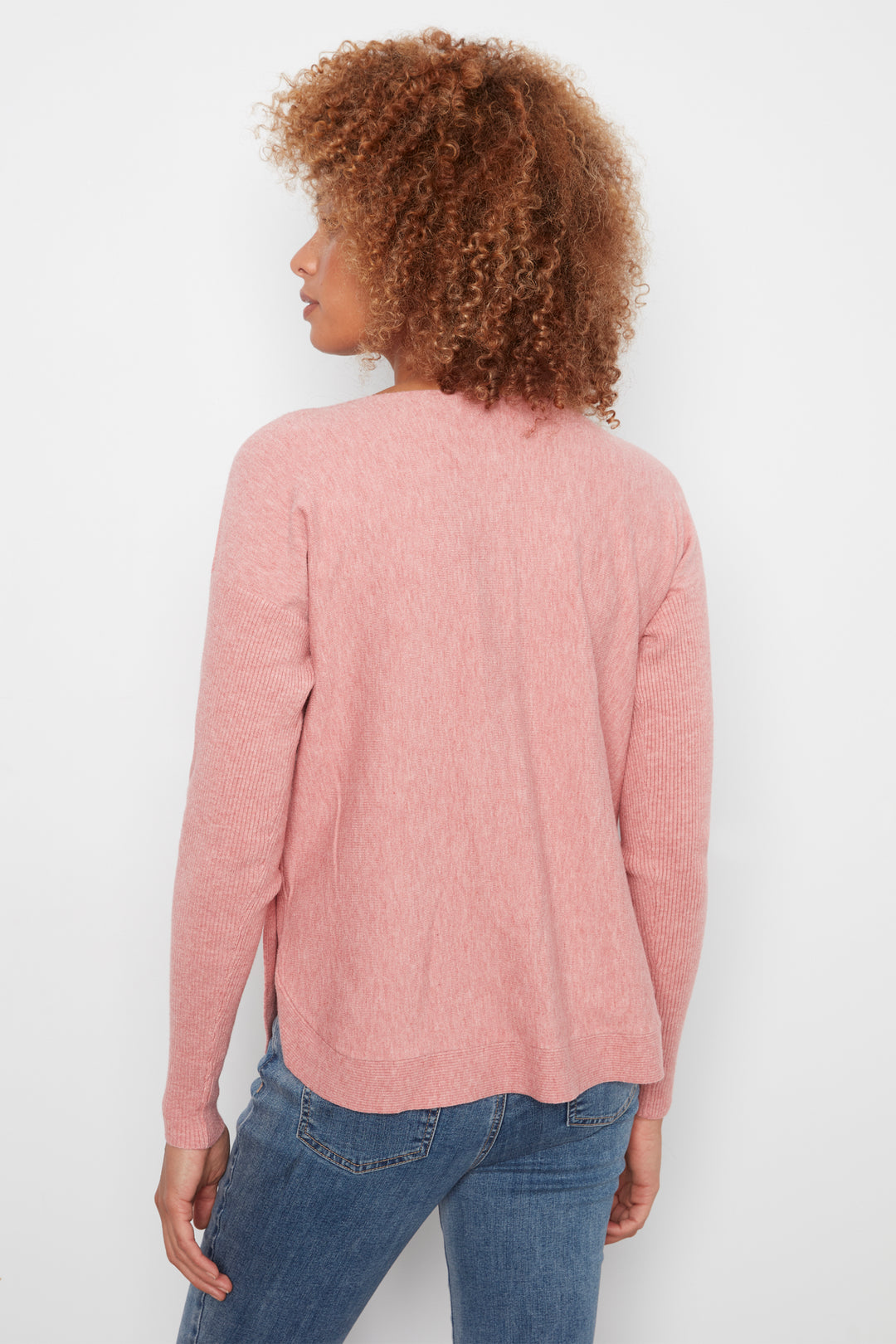 A woman with curly brown hair is shown from the back wearing a dusty pink long-sleeve knit sweater and blue jeans against a plain white background.