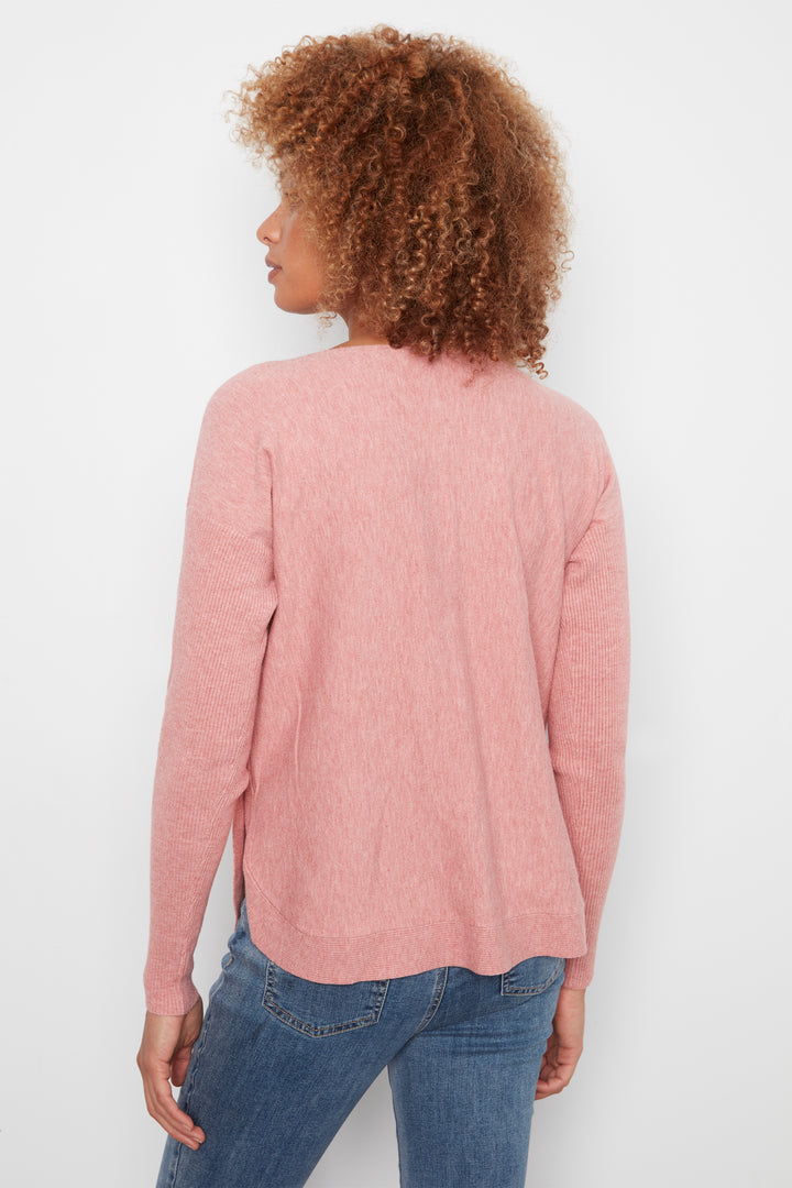 A woman with curly brown hair is shown from the back wearing a dusty pink long-sleeve knit sweater and blue jeans against a plain white background.