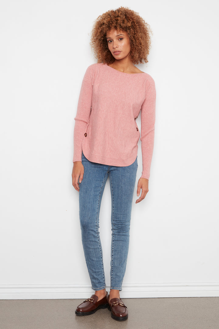 A woman with curly brown hair stands against a white wall wearing a dusty pink knit sweater, blue skinny jeans, and brown leather loafers.
