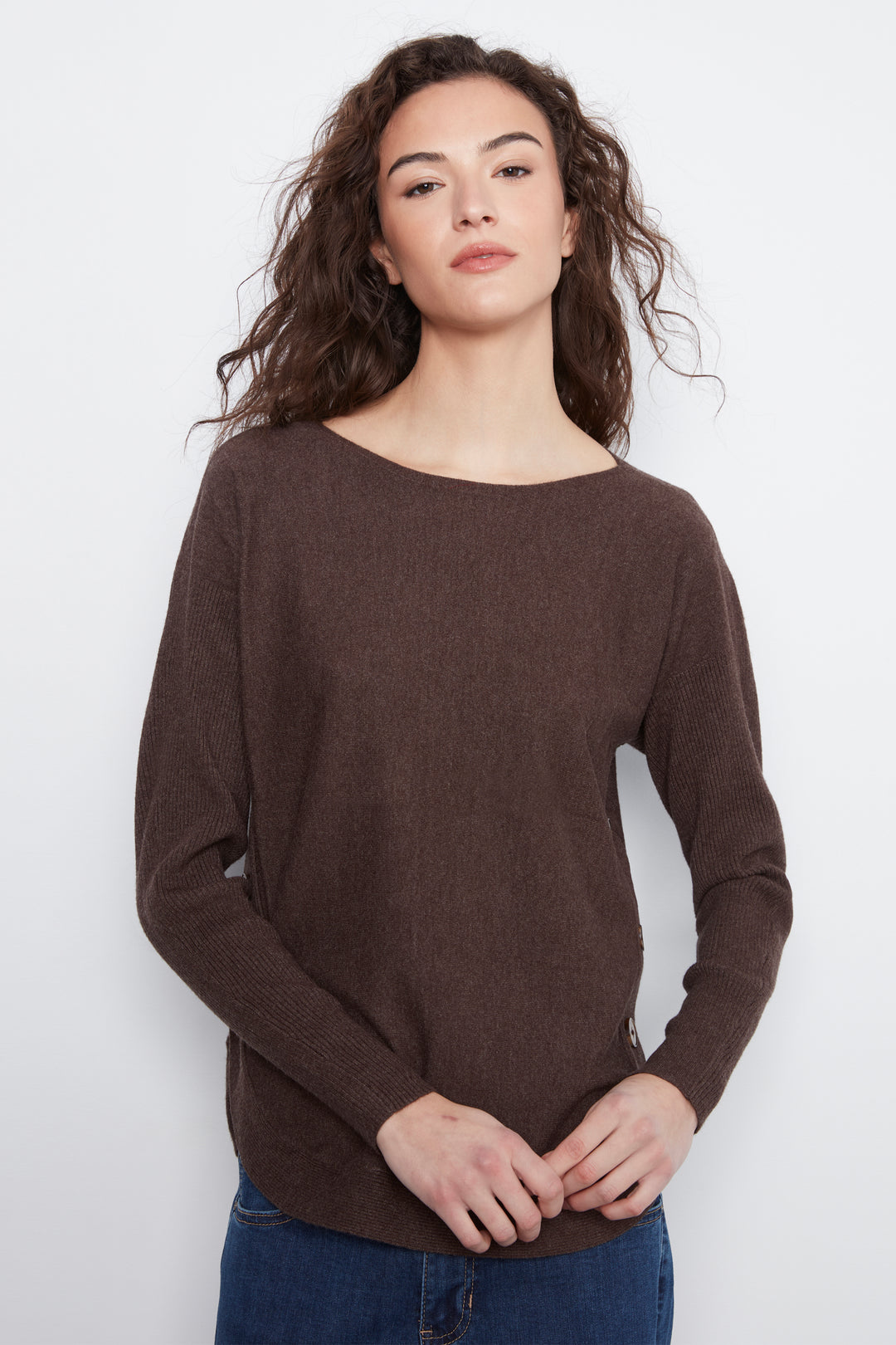 A woman with long, wavy brown hair wearing a dark brown boat-neck knit sweater and blue jeans, standing against a plain white studio background.