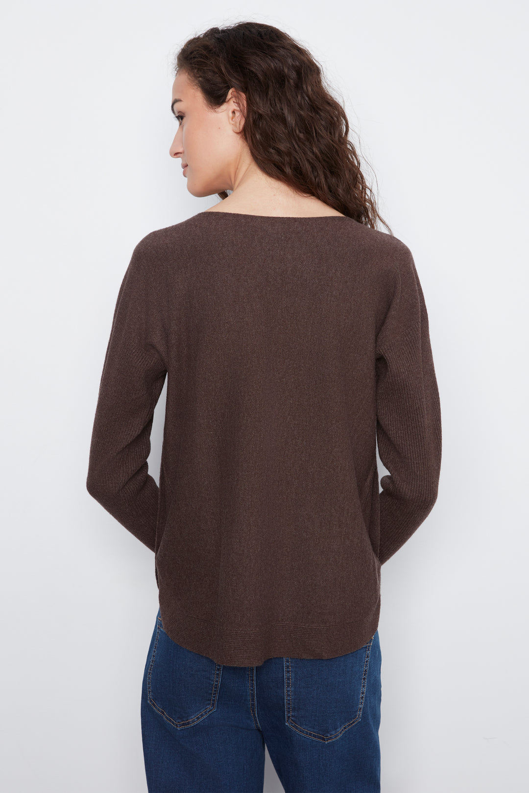 A woman shown from the back wearing a brown long-sleeve knit top and blue jeans against a plain light gray studio background.