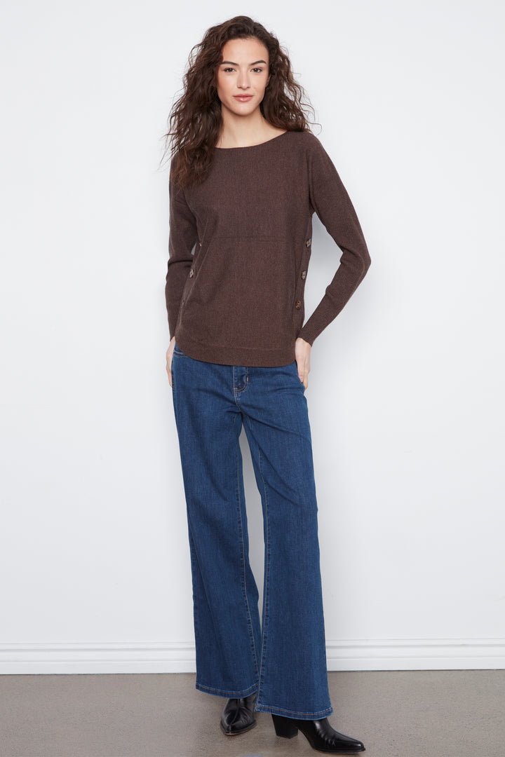 A woman modeling a brown long-sleeve knit top with a wide boat neckline and front kangaroo pocket, paired with blue wide-leg jeans and black shoes, standing against a white wall.