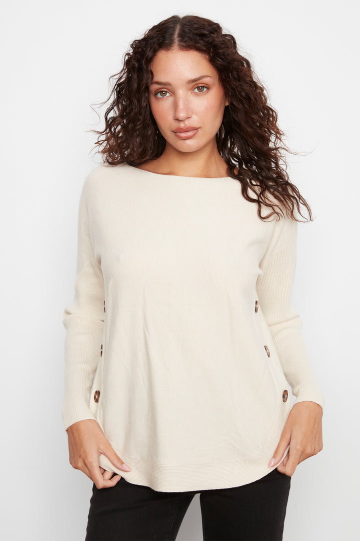 A woman with long, curly brown hair wearing a cream-colored long-sleeve knit top with brown buttons on the sides, paired with black pants, against a plain white studio background.