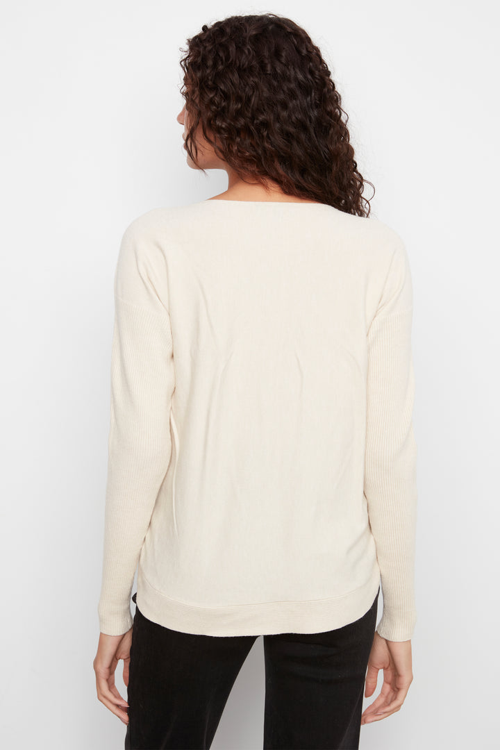 A woman with curly dark hair is shown from the back wearing a cream-colored long-sleeve top and black pants against a plain white background.