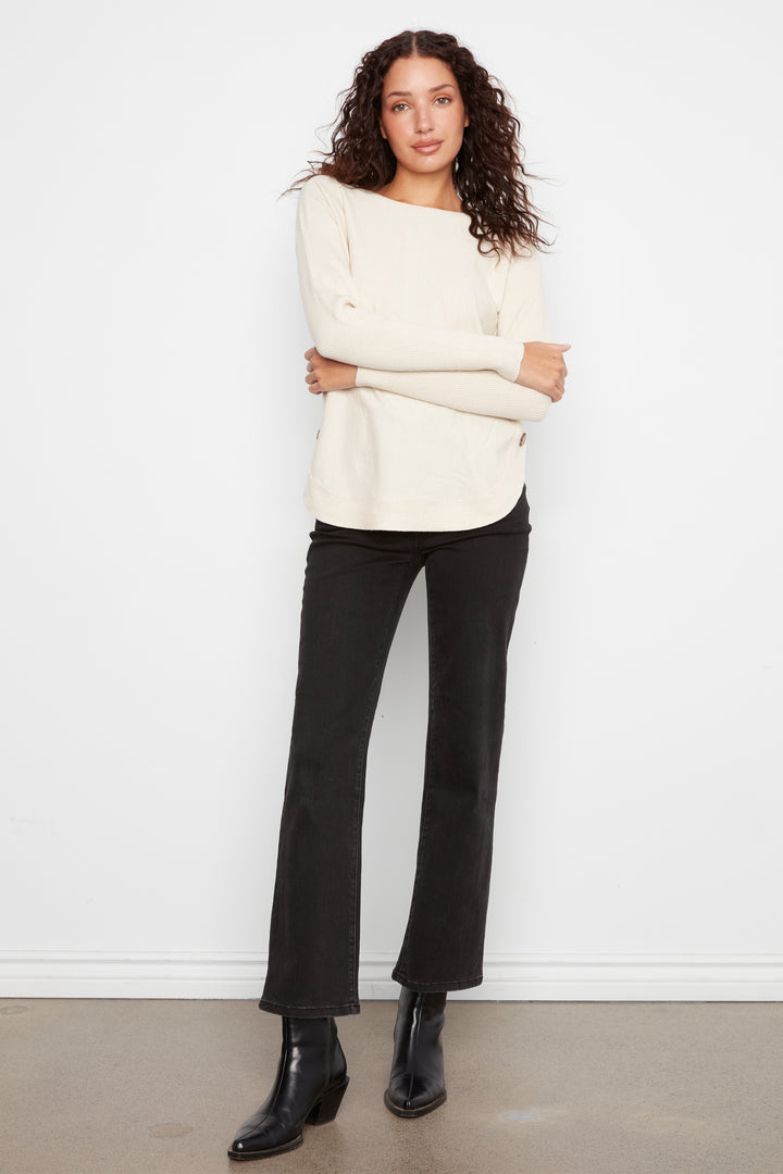 A woman modeling a cream off-the-shoulder knit sweater and black cropped flare pants with black ankle boots, standing against a white wall.