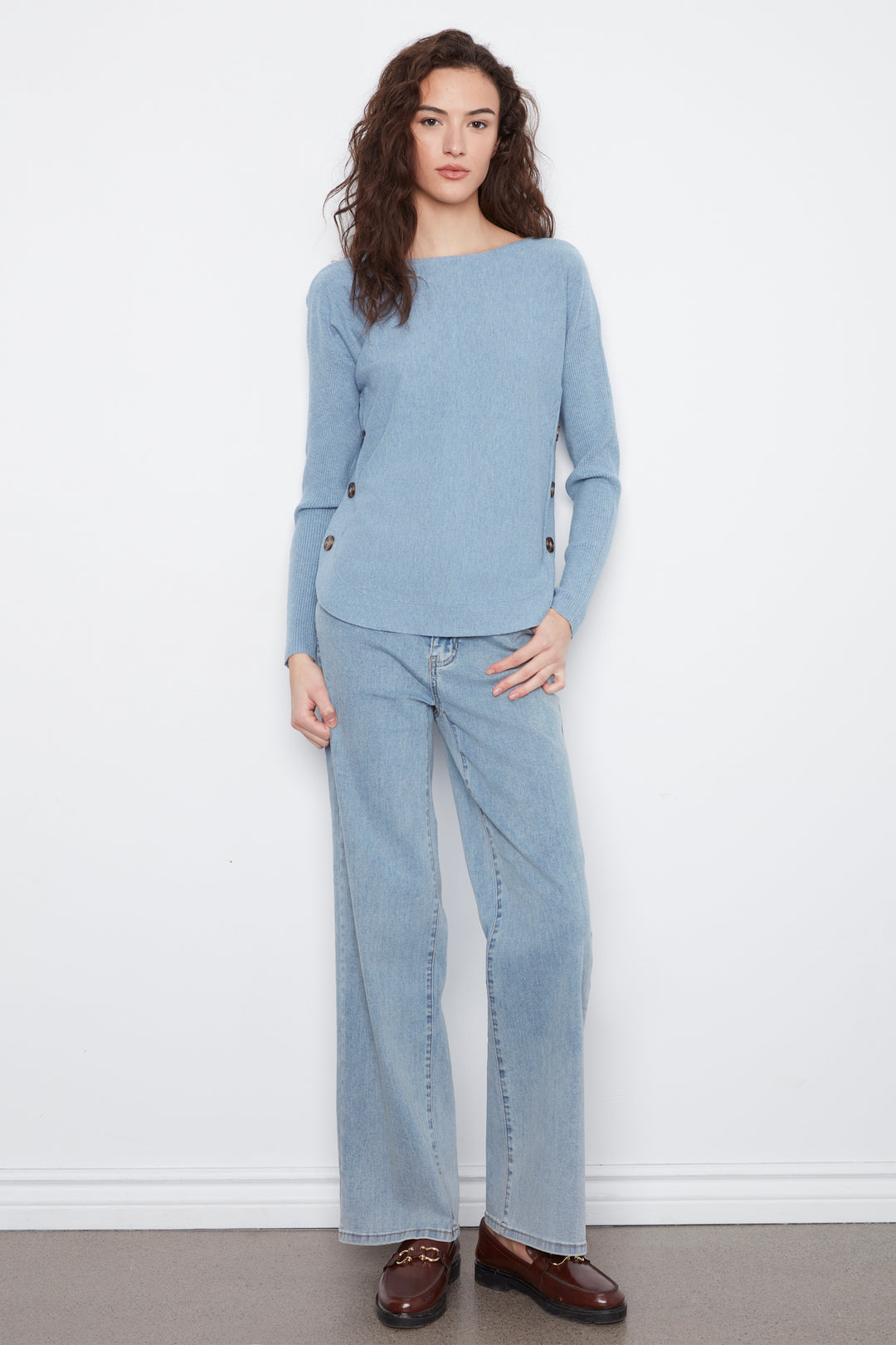 A woman modeling a light blue knit sweater and matching wide-leg jeans against a plain white wall, wearing brown leather loafers.