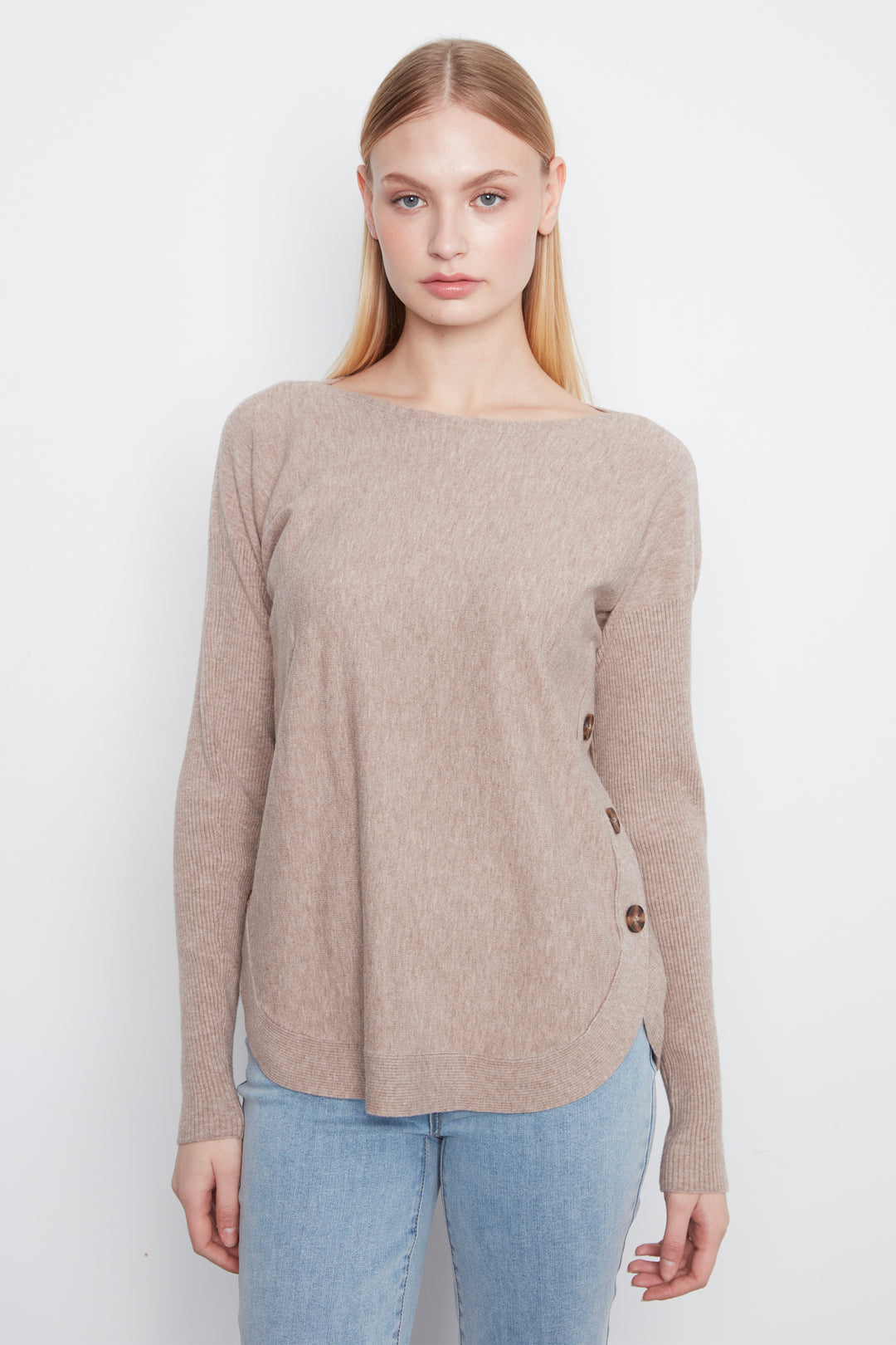 A woman modeling a light taupe knit sweater with a boat neckline and decorative brown buttons on the side, paired with light blue jeans against a plain white background.