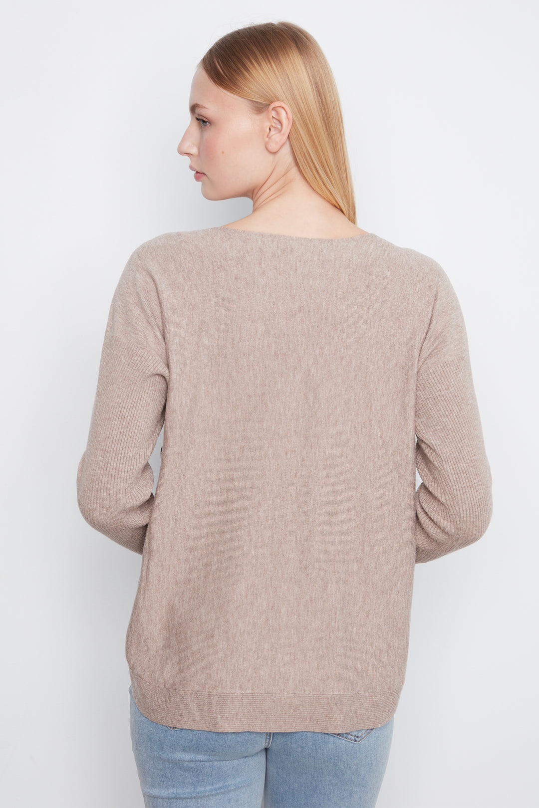 A woman shown from the back wearing a light beige knit sweater and light blue jeans against a plain white studio background.