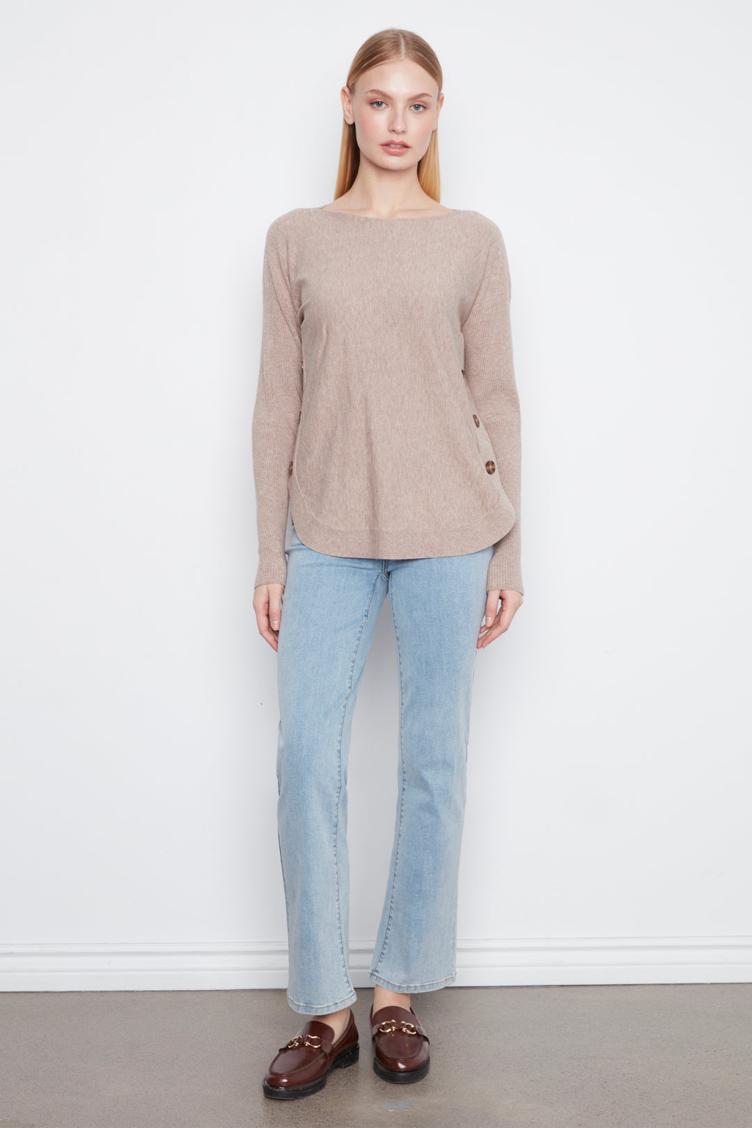 A woman stands against a plain white wall wearing a light taupe-beige long-sleeve knit sweater, light-wash blue jeans, and dark brown leather loafers.