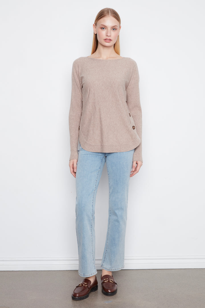 A woman stands against a plain white wall wearing a light taupe-beige long-sleeve knit sweater, light-wash blue jeans, and dark brown leather loafers.