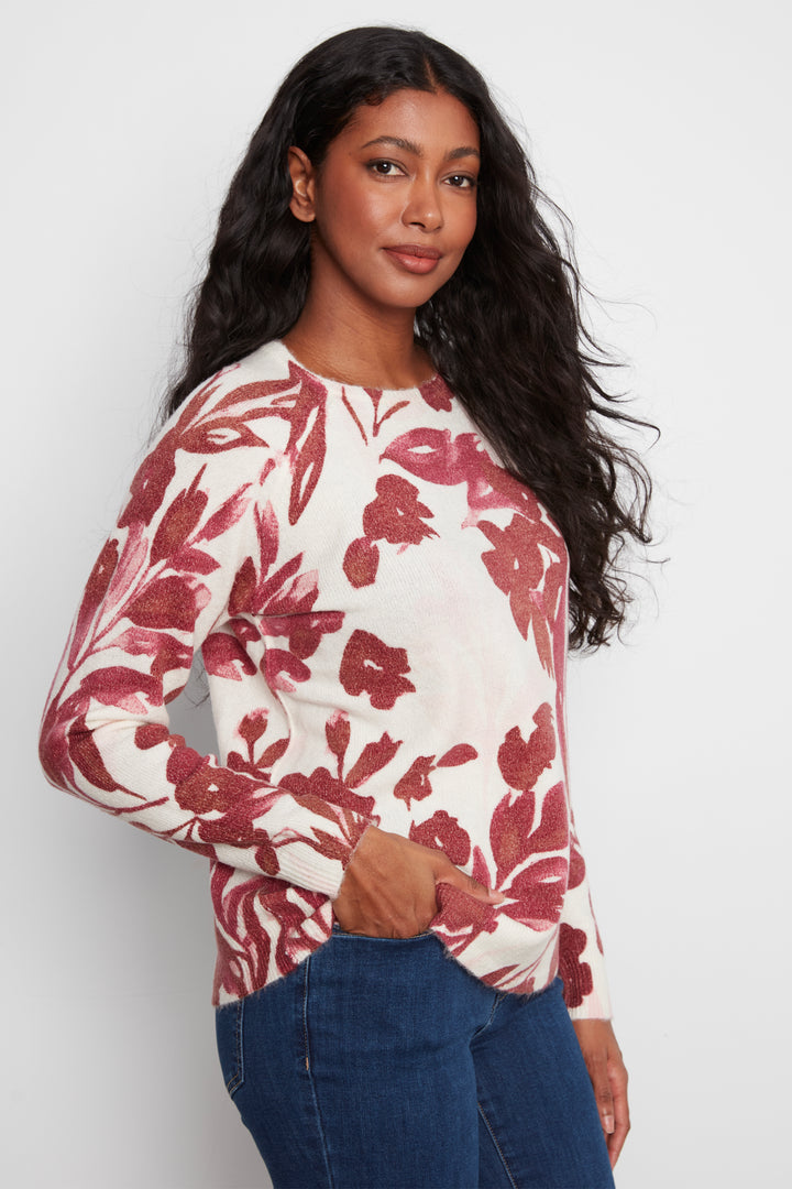 A woman modeling a long-sleeve knit top with a red and burgundy floral pattern on a cream background, paired with blue jeans, against a light gray studio background.