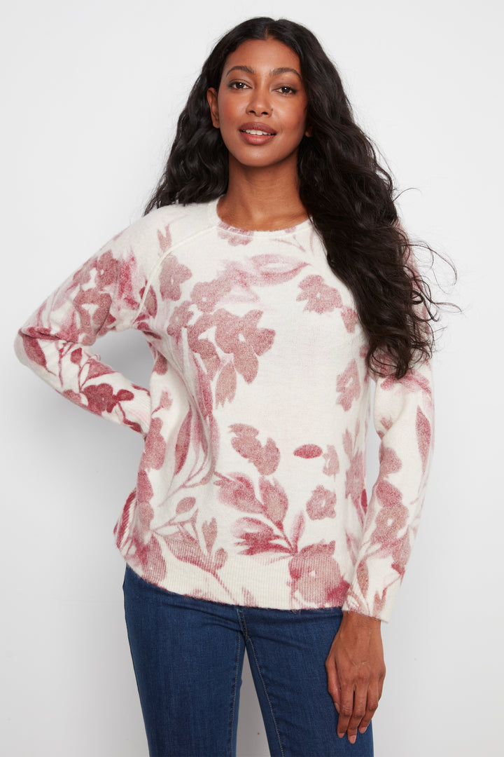 A woman modeling a cream-colored long-sleeve knit top with a dusty rose floral pattern, paired with blue jeans, against a plain light gray background.