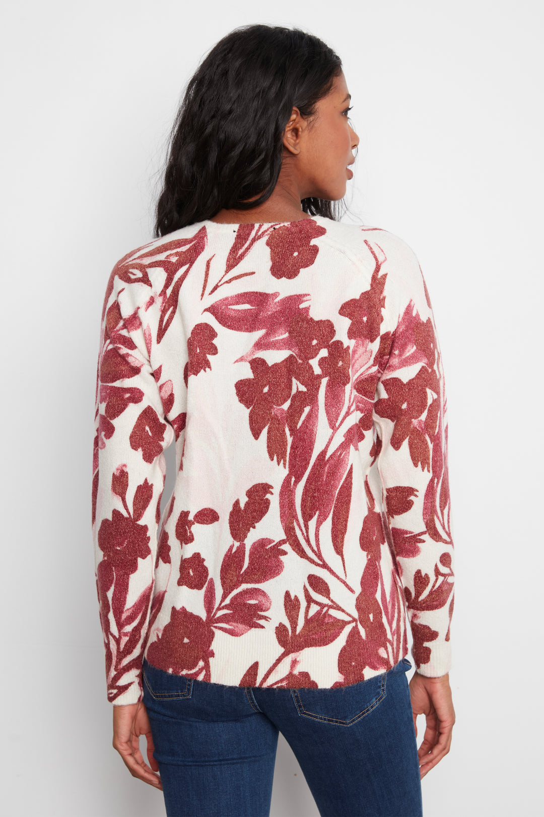 A woman shown from the back wearing a cream-colored long-sleeve top with a red floral pattern, paired with blue jeans, against a plain light gray background.