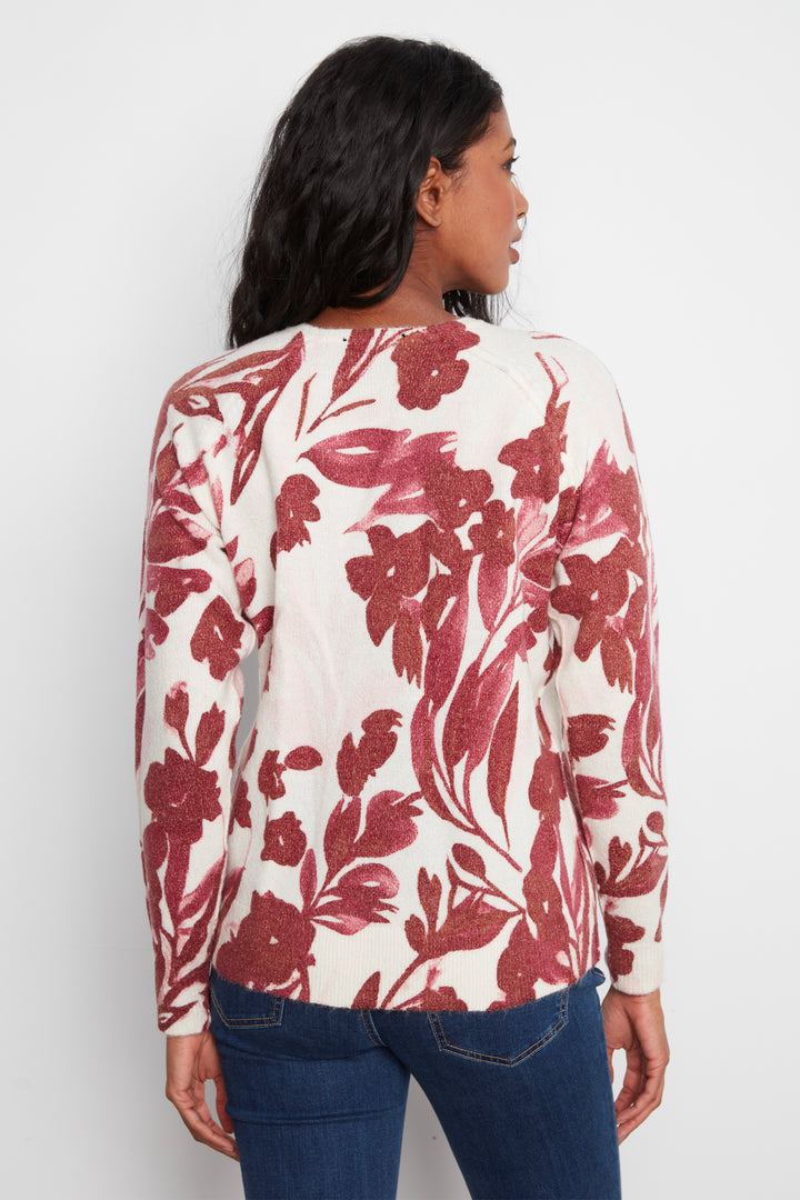 A woman shown from the back wearing a cream-colored long-sleeve top with a red floral pattern, paired with blue jeans, against a plain light gray background.