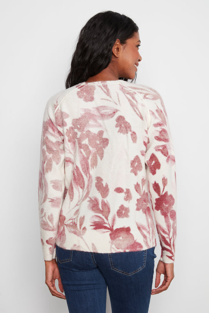 A woman viewed from the back wearing a cream-colored long-sleeve top with a dusty rose floral pattern, paired with dark blue jeans, against a plain light gray background.