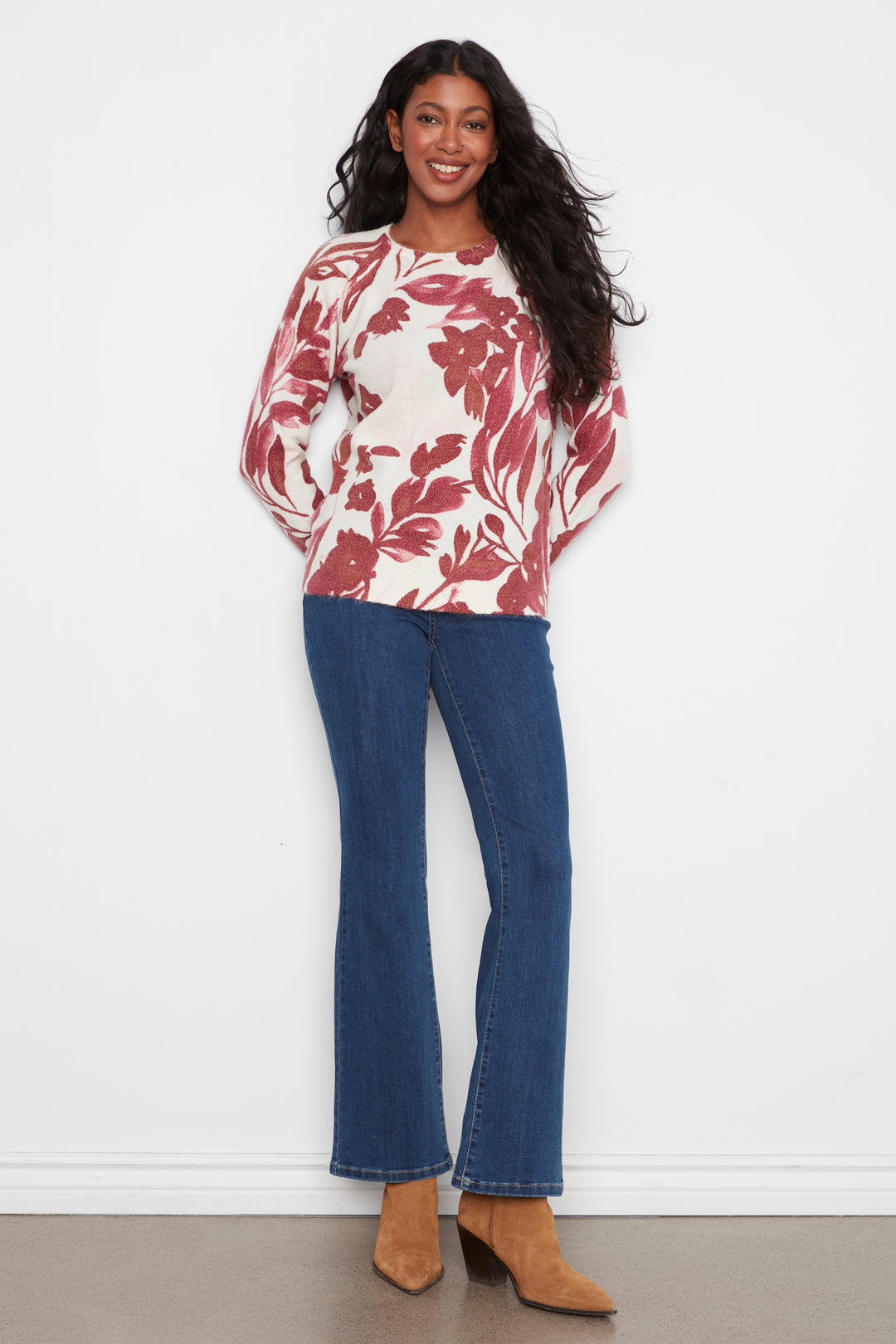 A woman modeling a long-sleeve crewneck top with a red and white floral pattern, paired with blue flare jeans and tan ankle boots, standing against a white wall.