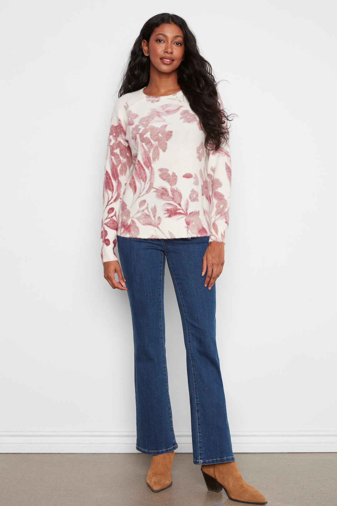A woman modeling a cream long-sleeve crewneck sweater with a dusty pink floral pattern, paired with blue flare jeans and brown suede ankle boots, standing against a white wall.