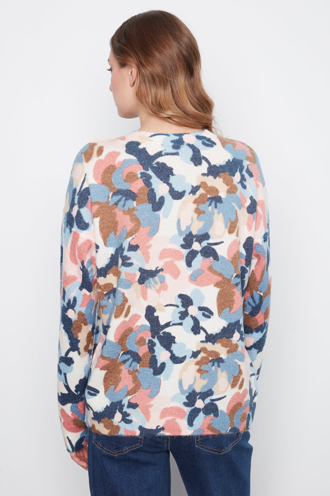 A woman viewed from the back wearing a long-sleeve knit top with a multicolor abstract floral pattern in blue, pink, and brown on a cream background, paired with dark blue jeans against a plain white studio background.