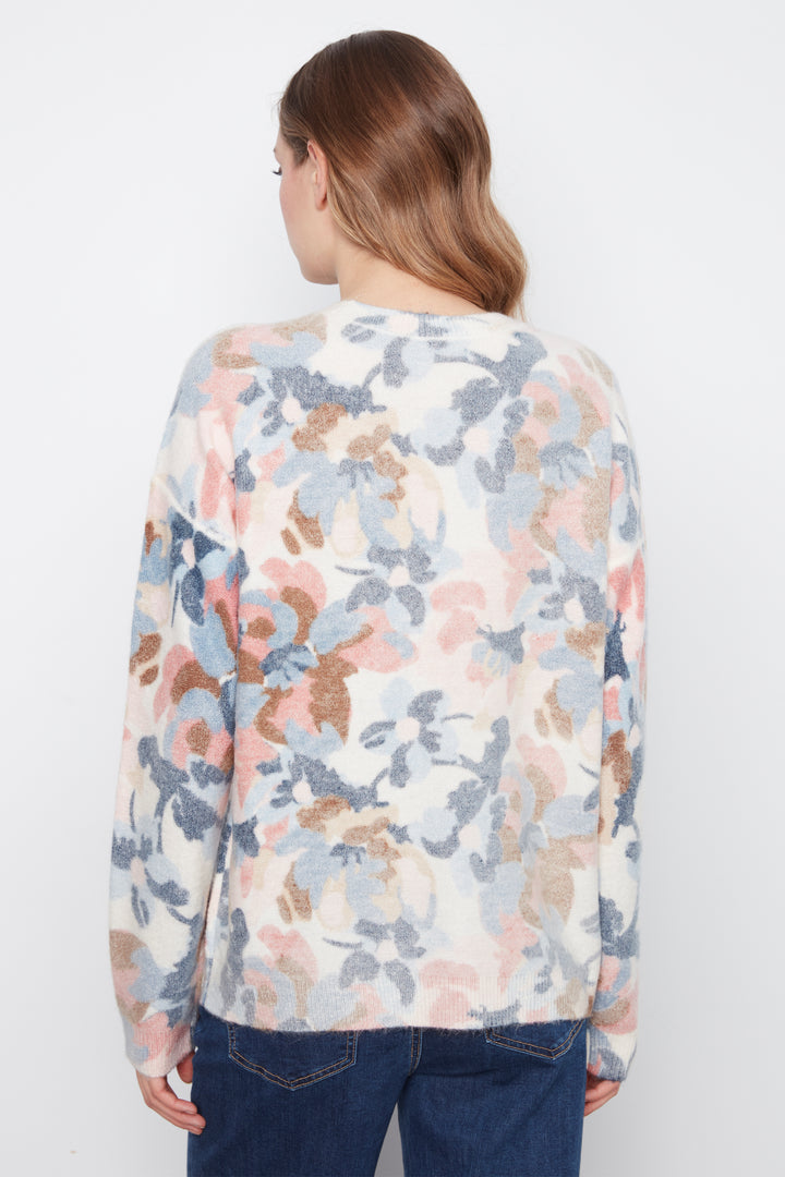 A woman viewed from behind wearing a cream-colored knit sweater with a soft, painterly floral pattern in dusty pink, slate blue, and brown, paired with dark blue jeans against a plain white background.