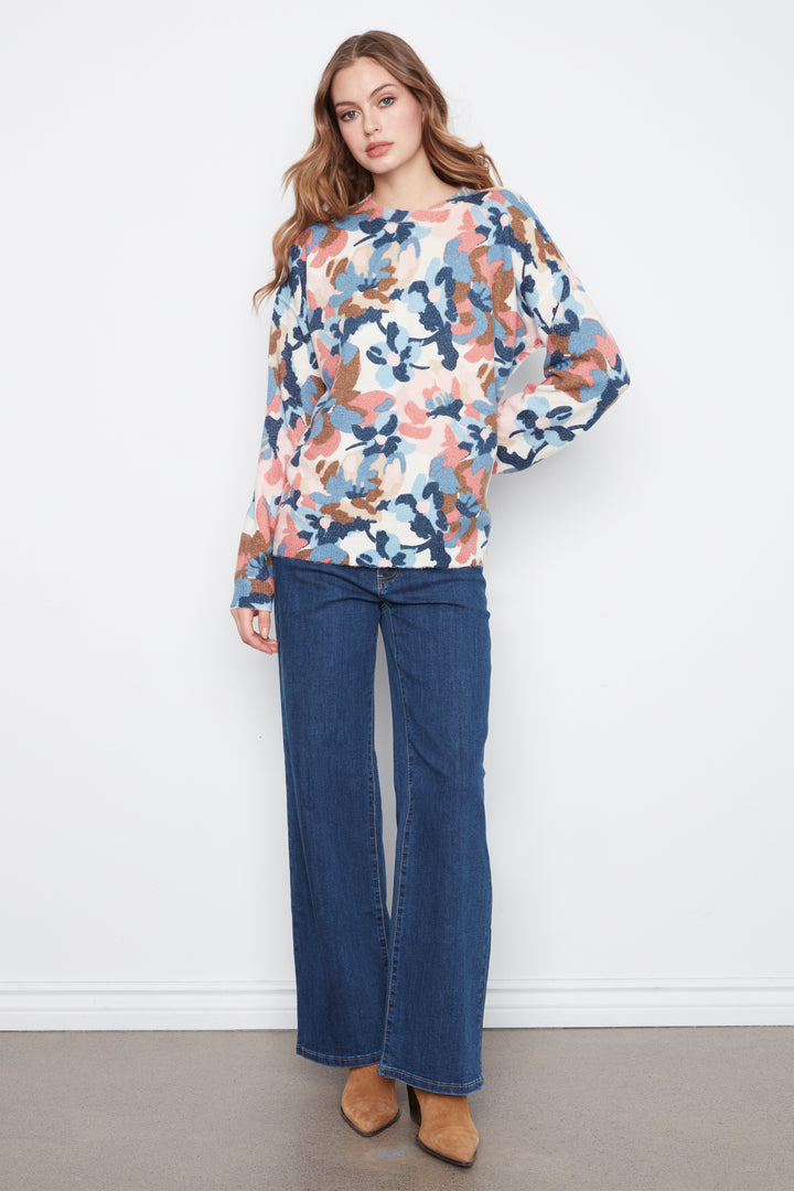 A woman modeling a long-sleeve sheer blouse with a blue, pink, and brown floral pattern, paired with dark blue wide-leg jeans and tan pointed-toe flats, standing against a white wall.