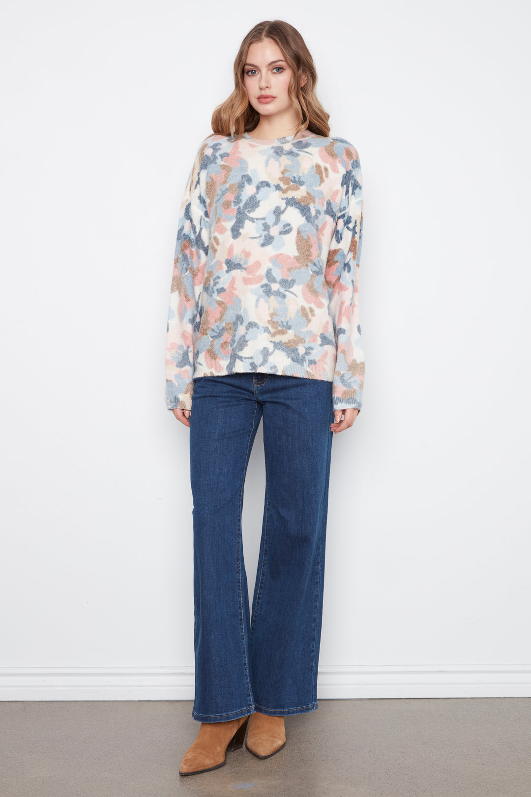 A woman stands against a white wall wearing a long-sleeve floral top with blue, pink, and beige tones, paired with dark blue wide-leg jeans and tan ankle boots.
