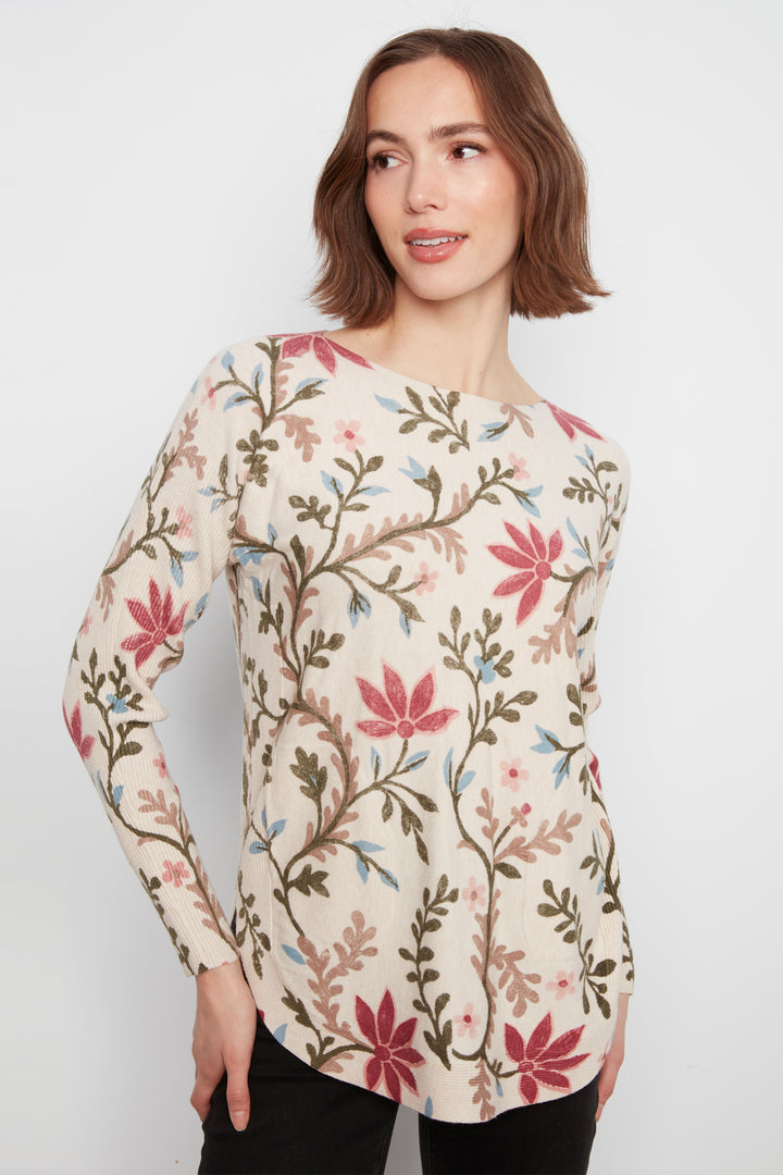A woman wearing a long-sleeve cream-colored top with a colorful floral and botanical pattern in pink, green, and brown, paired with black pants, set against a plain light gray background.