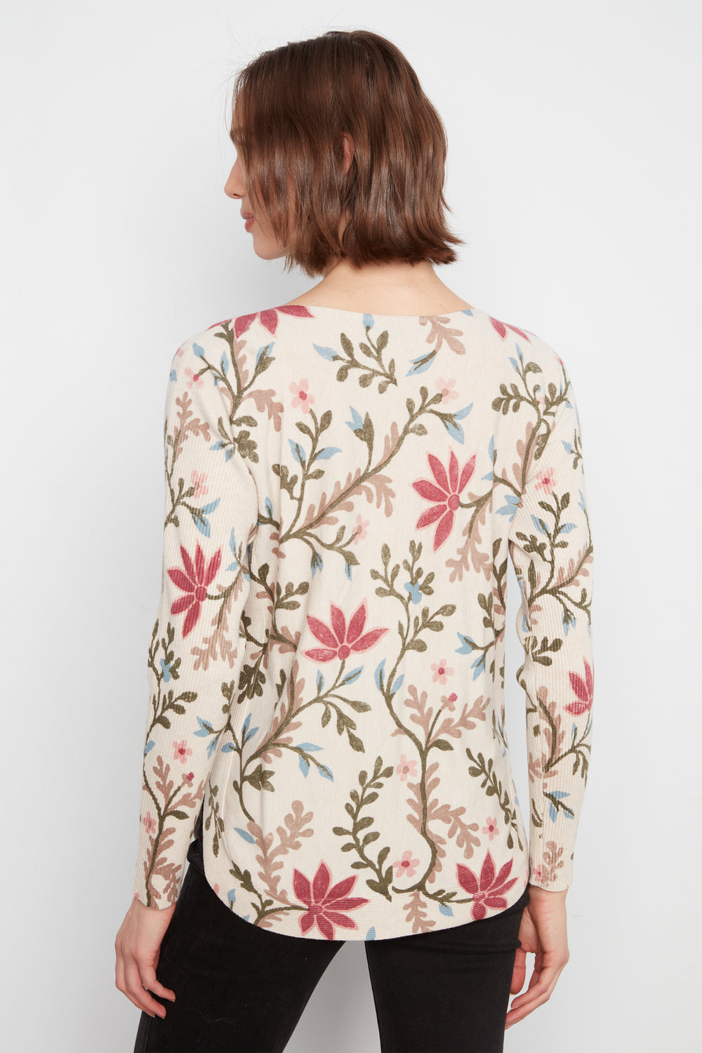 A woman shown from the back wearing a long-sleeve cream-colored top with a dense floral and botanical pattern in pink, green, and brown, paired with black pants against a plain white background.