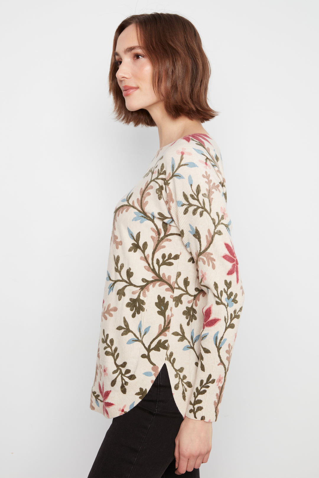 A woman shown in side profile wearing a cream-colored long-sleeve top with an all-over botanical branch and leaf pattern in olive green, dusty pink, and light blue, paired with black pants, against a plain light gray background.