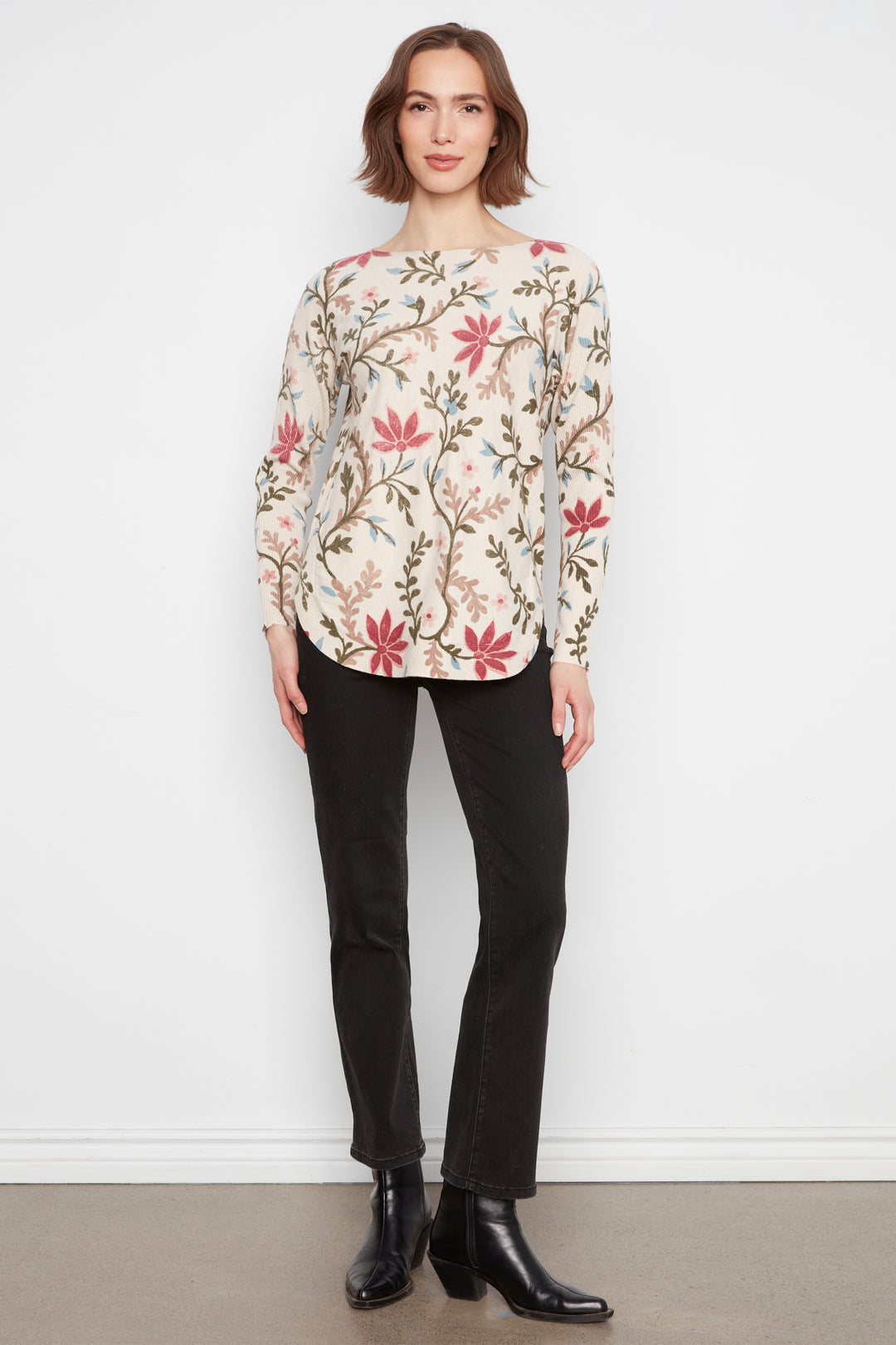 A woman stands against a white wall wearing a cream-colored long-sleeve blouse with a red and green floral pattern, paired with black pants and black ankle boots.