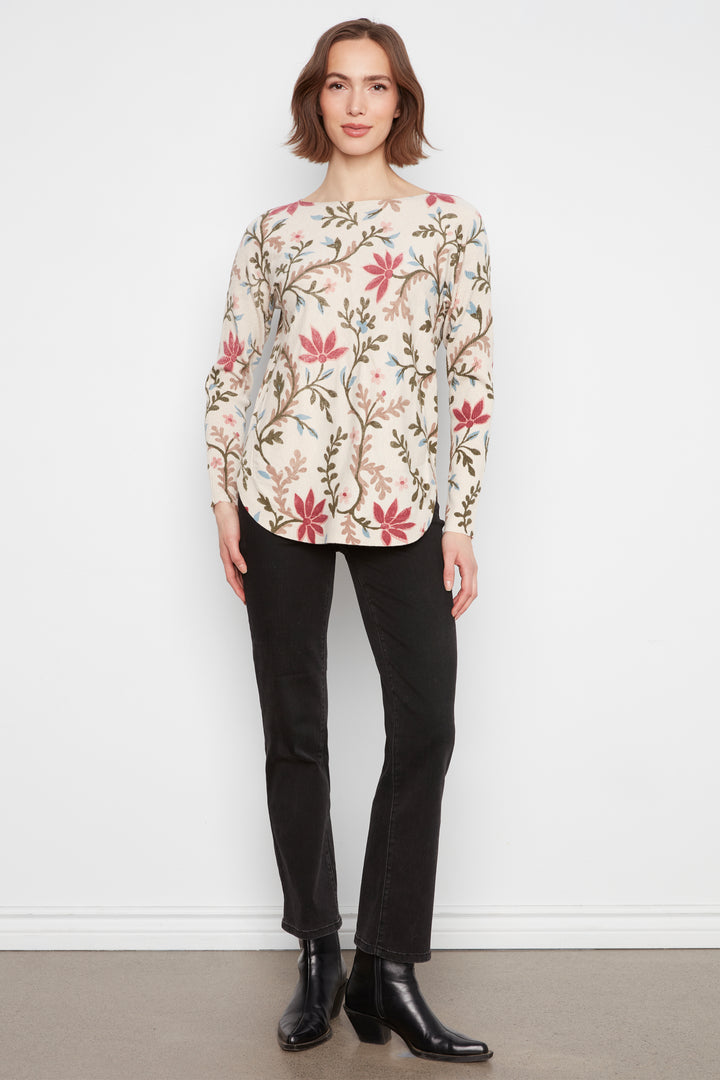 A woman stands against a white wall wearing a cream-colored long-sleeve blouse with a red and green floral pattern, paired with black pants and black ankle boots.