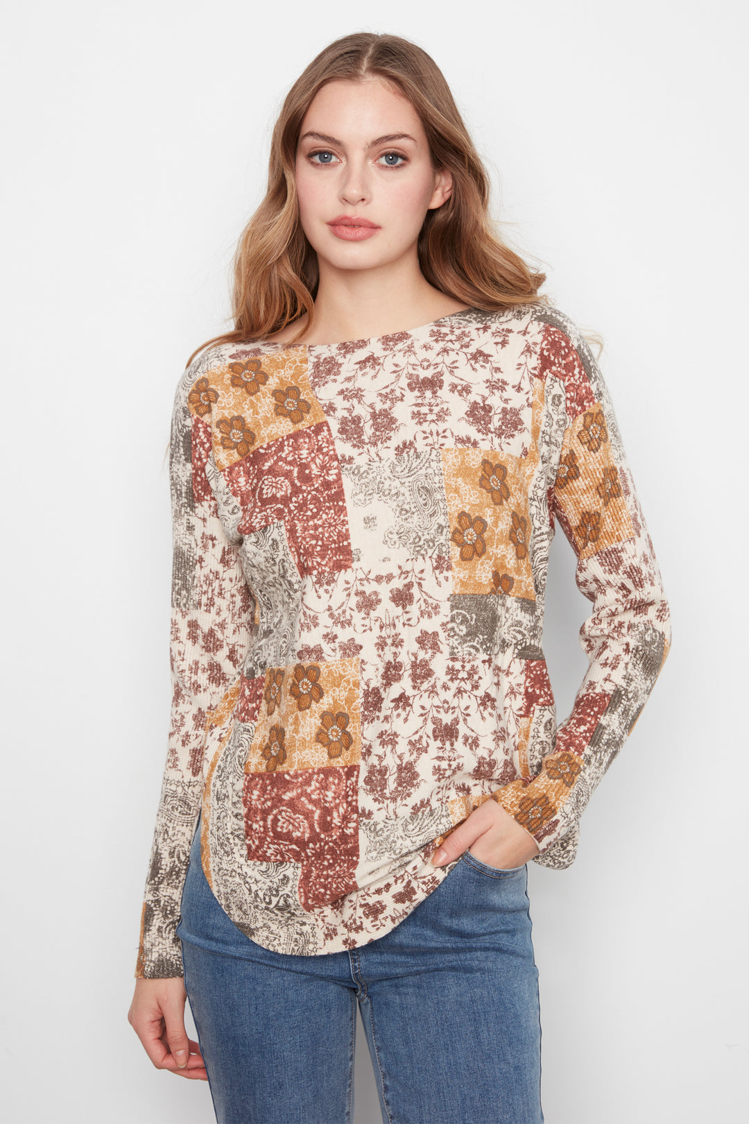 A woman modeling a long-sleeve patchwork blouse with a bohemian floral print in rust, mustard, and beige tones, paired with blue jeans against a plain white studio background.