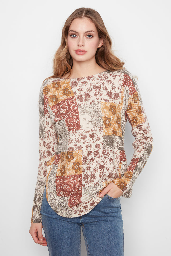 A woman modeling a long-sleeve patchwork blouse with a bohemian floral print in rust, mustard, and beige tones, paired with blue jeans against a plain white studio background.