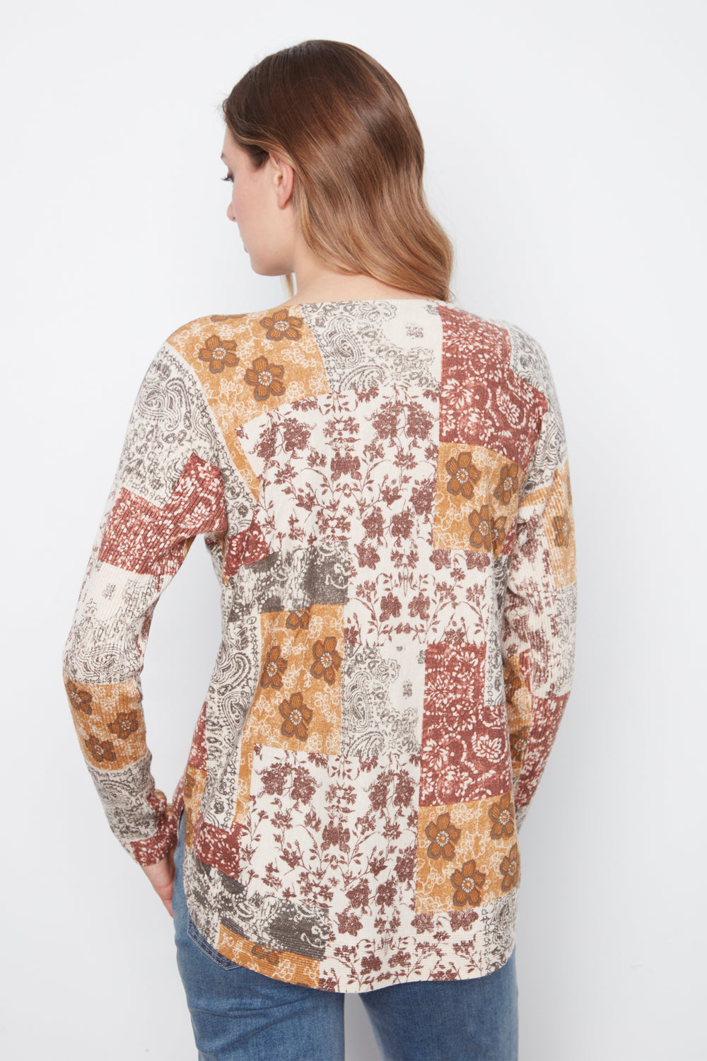 A woman shown from the back wearing a long-sleeve patchwork top with a floral and paisley print in rust red, mustard yellow, and beige, paired with blue jeans against a plain white background.