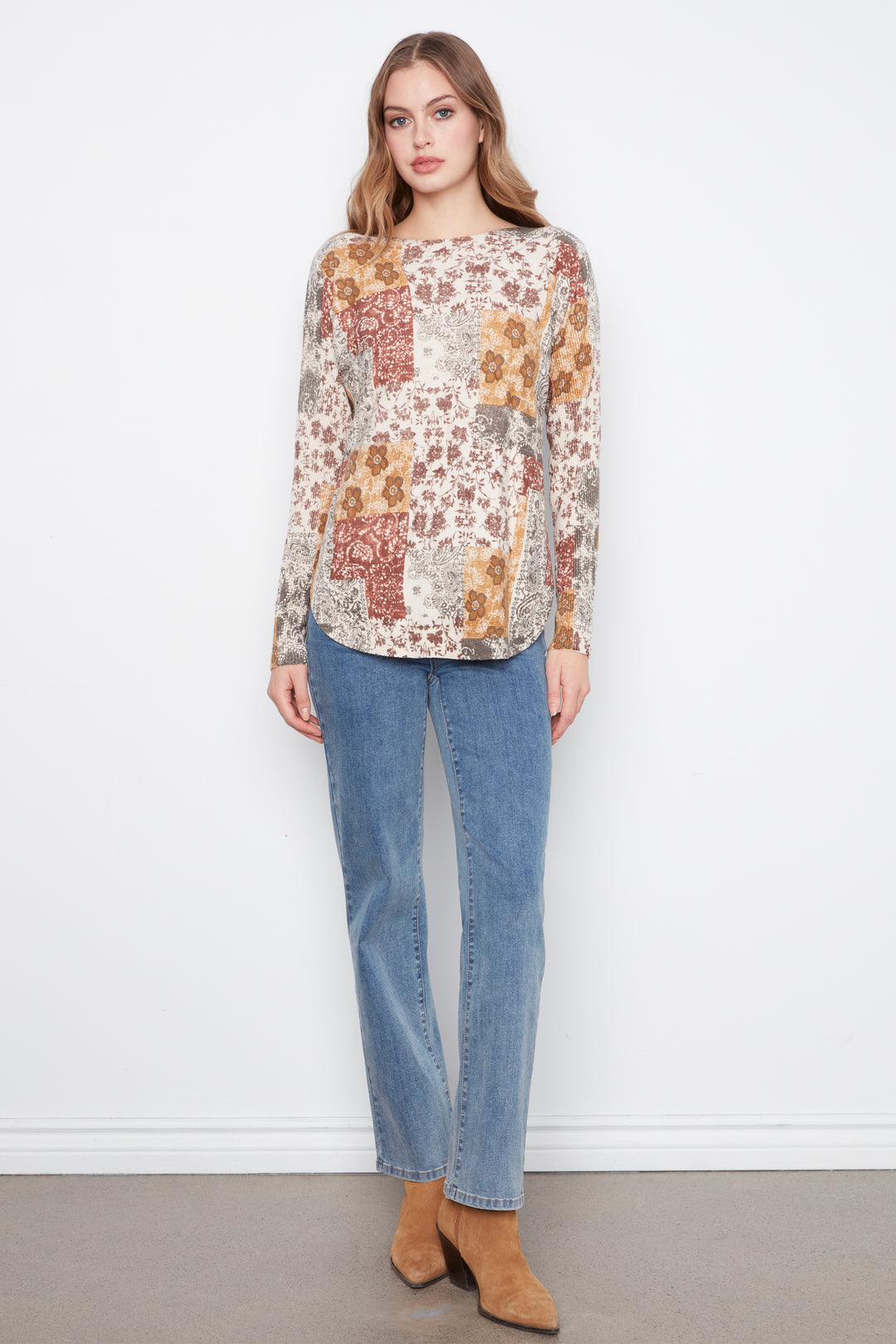 A woman modeling a long-sleeve patchwork floral top with blue jeans and tan ankle boots, standing against a white wall.