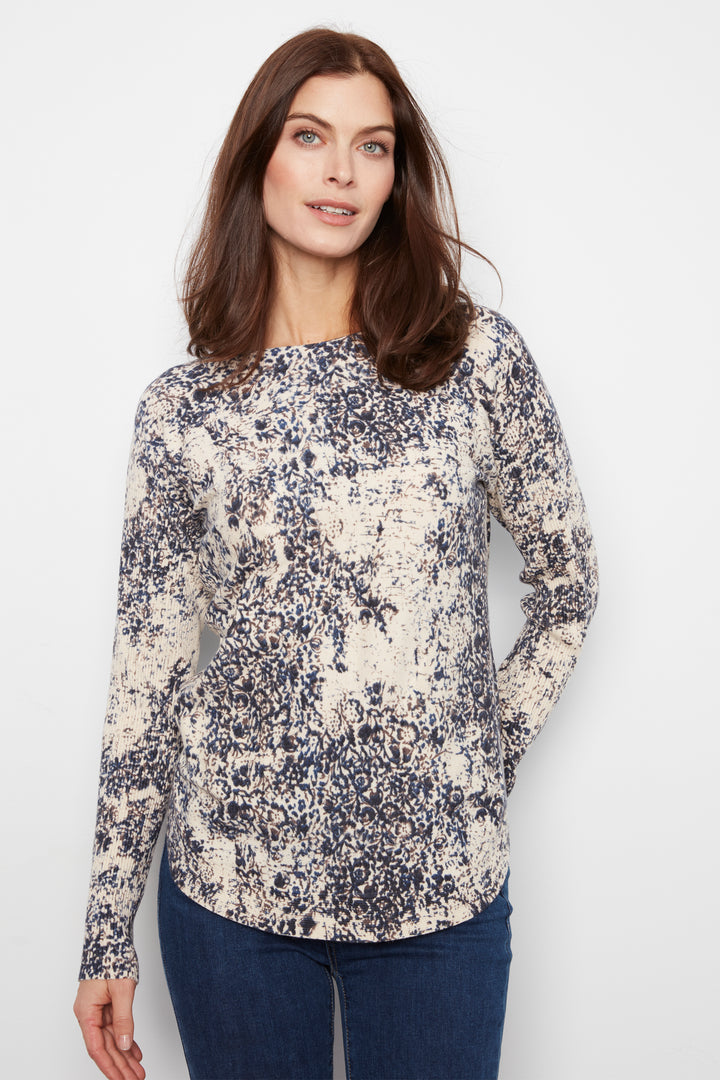 A woman modeling a long-sleeve knit top with a navy and cream abstract watercolor pattern, paired with dark blue jeans, against a plain light gray studio background.