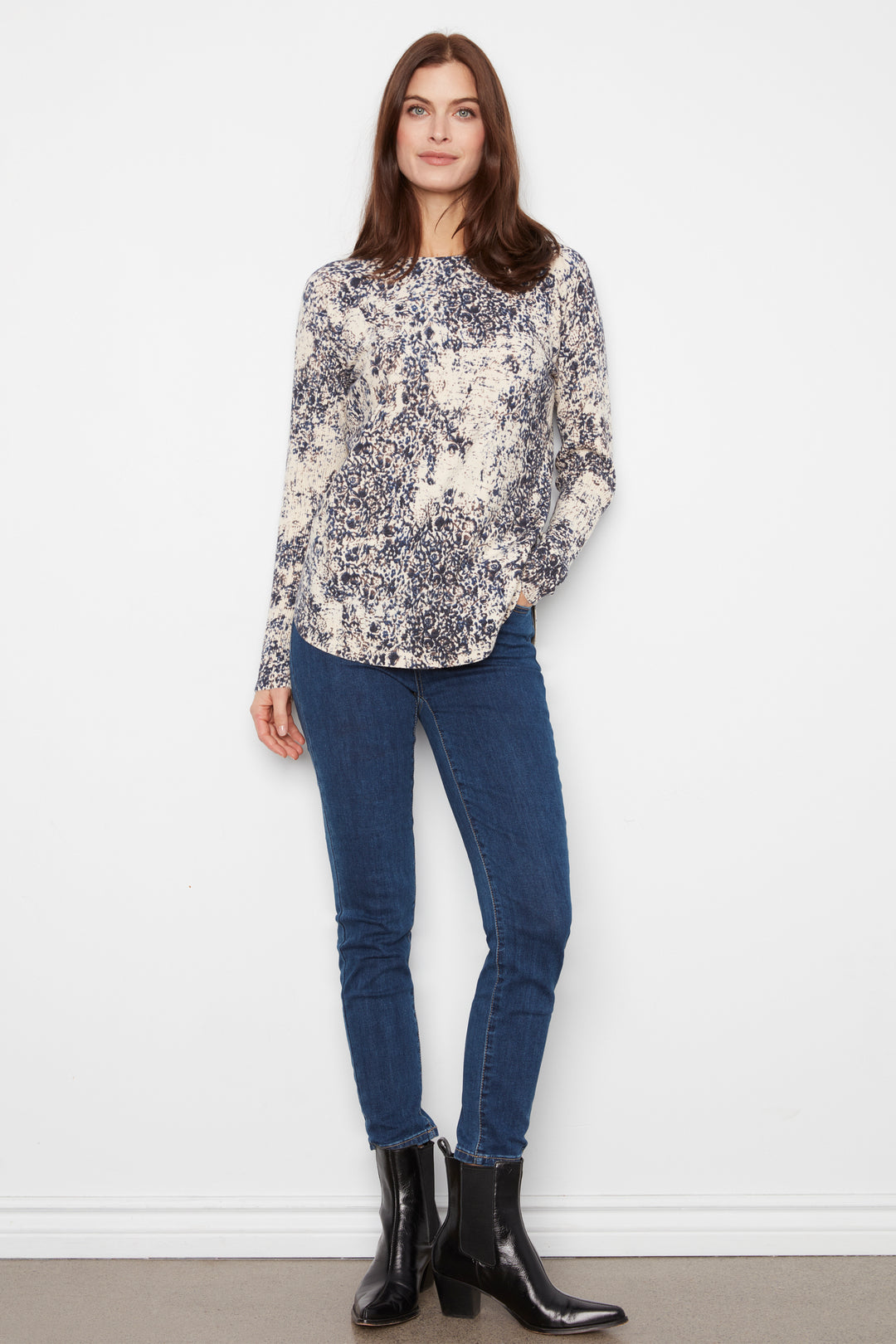 A woman modeling a long-sleeve blouse with a blue and beige floral pattern, paired with dark blue skinny jeans and black ankle boots, standing against a plain white wall.