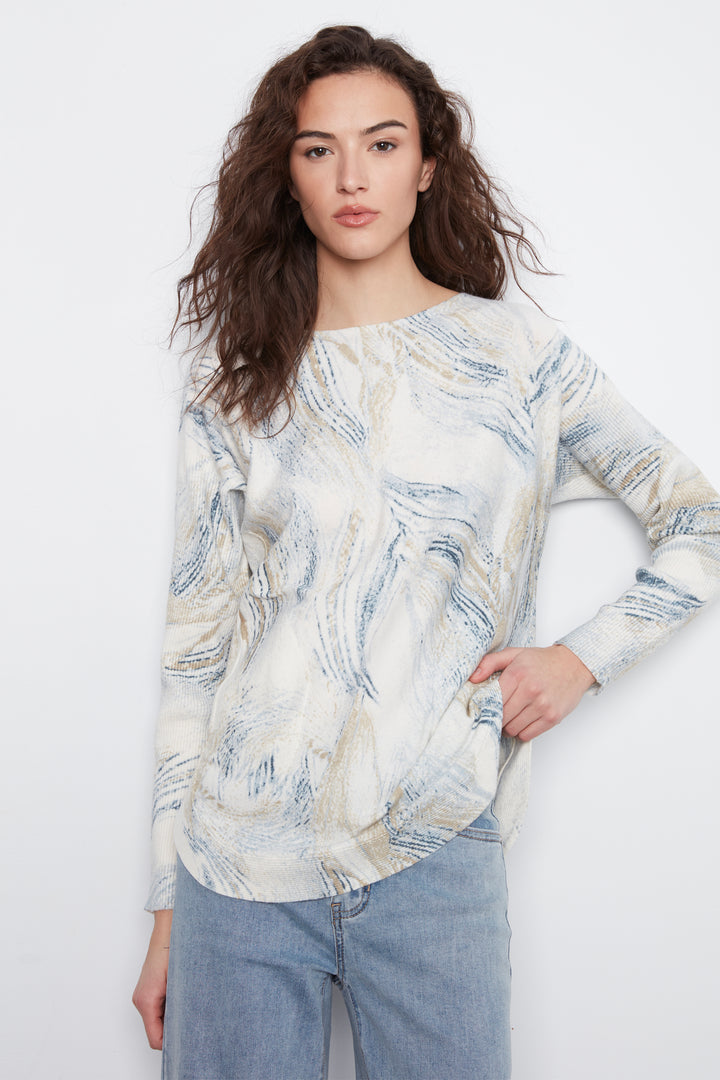 A woman modeling a long-sleeve top with an abstract blue, beige, and white marbled pattern, paired with light blue jeans, against a plain white background.