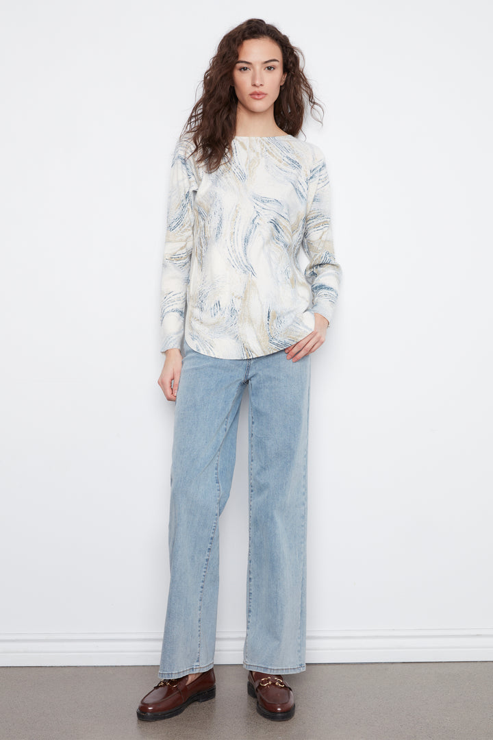 A woman stands against a white wall wearing a cream and light blue abstract-patterned long-sleeve top, light-wash wide-leg jeans, and brown leather loafers.